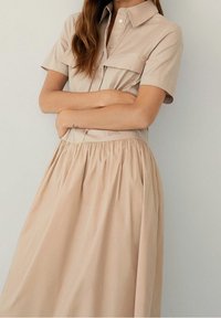 Abito camicia color sabbia con.collo a camicia, maniche corte e due tasche sul petto, caratterizzato da una gonna arricciata e una texture di tessuto liscia.