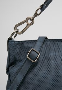 Gros plan sur un sac en cuir perforé bleu avec une bandoulière réglable et un fermoir en métal gravé "SURFREY" sur fond gris uni.