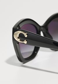 Lunettes de soleil noires avec une forme angulaire audacieuse. Elles présentent des verres dégradés violets et un accent doré sur la branche. Fini en plastique lisse.