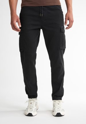 Mann trägt schwarze Cargo-Jogginghosen mit Kordelzugbund und weiße Turnschuhe, steht vor einem weißen Hintergrund.