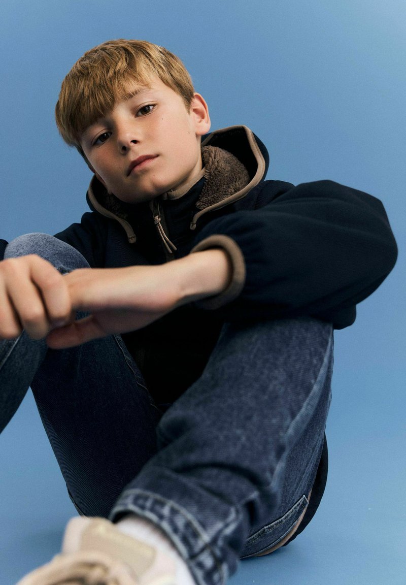 Jeune garçon aux cheveux châtain clair assis en tailleur, portant une veste sombre et un jean bleu sur un fond bleu uni.