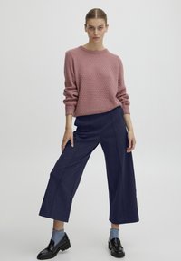 Roze gestructureerde gebreide trui met een ronde halslijn, gecombineerd met marineblauwe wijde broekspijpen. Model draagt donkere schoenen en lichtblauwe sokken.