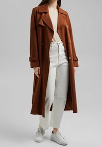 Cappotto trench marrone con un ampio colletto, bottoni doppi, e maniche arrotolate, abbinato a jeans bianchi e sneakers bianche.
