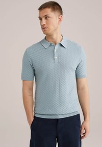 Svetlo modra polo majica s kratkimi rokavi z teksturiranim, mrežastim vzorcem in klasičnim ovratnikom. Ima tri gumbe ter rebraste rokave in spodnji rob.