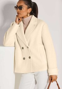 Vrouw in een crèmekleurige dubbelzijdige jas, beige turtleneck, zonnebril, gouden oorbellen, met een bruine handtas, poseert tegen een lichte achtergrond.
