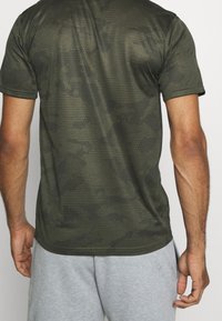 Krátký rukáv atletického trička v zeleném maskáčovém vzoru, s texturovaným vzorem ve tvaru včelí plástve, vyrobeného z lehkého materiálu a s přiléhavým střihem.