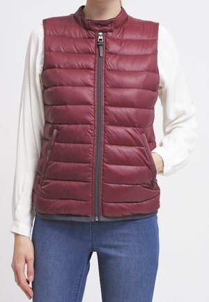 Frau trägt eine ärmellose, gesteppte bordeauxrote Weste, die über einer langärmligen weißen Bluse und blauen Jeans hochgeschlossen ist, die Hände teils in den Westentaschen.