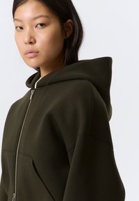 Νεαρή γυναίκα με ίσια σκούρα μαλλιά, φοράει σκούρα πράσινη φούτερ με φερμουάρ και σηκωμένη κουκούλα, κοιτάζοντας ελαφρώς εκτός κάδρου με φόντο λευκό.