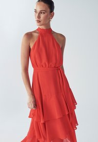 Red halter-neck dress made of lightweight fabric, featuring tiered layers and a fitted waist with a tied belt for a flowing silhouette.