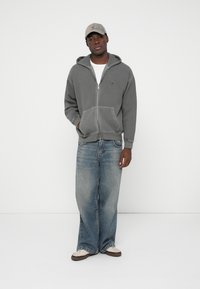 Grauer Reißverschluss-Hoodie mit Kängurutasche, kombiniert mit hellblauen Jeans und beigen Sneakers; trägt eine hellfarbene Kappe.