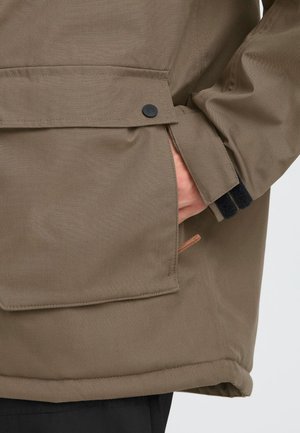 Abrigo de tela marrón texturizada con un bolsillo lateral, cierre de botón negro y puños negros en contraste. Sutil acento de diseño con un ribete color beige.