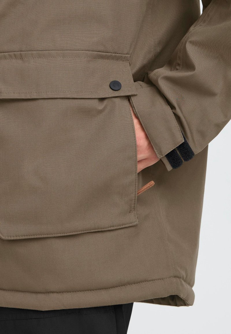 Rudas, tekstūruotas audinio paltas su šonine kišenę, juodų mygtukų užsegimu ir kontrastingais juodais rankogaliais. Subtilus dizaino akcentas su smėlio spalvos apvadu.