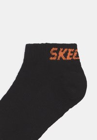 Juoda kulkšninių kojinių su tekstūruotu audiniu ir oranžine "SKEL" logotipu, įaustintu į kraštą. Paprastas dizainas, be raštų ar papildomų akcentų.