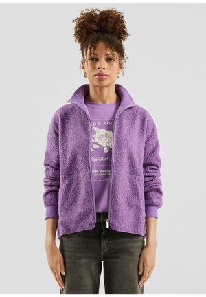 Frau mit zusammengebundenen lockigen Haaren trägt eine violette Fleecejacke über einem grafischen T-Shirt und dunklen Jeans vor einem schlichten, hellen Hintergrund.