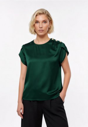 Femme aux cheveux blonds courts portant un chemisier en satin vert foncé avec des boutons dorés sur l'épaule et un pantalon noir, debout devant un fond blanc.