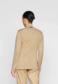 Μακρυμάνικο, μπεζ μπλουζάκι με ραβδωτή υφή και διπλωμένο γιακά, με μικρό λογότυπο στην πλάτη και λεία ραφές.