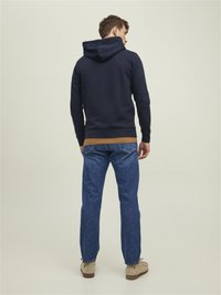 Pulover cu glugă de culoare bleumarin, cu un buzunar tip cangur și tiv contrastant maro, purtat cu blugi blue denim și pantofi bej, prezentat din spate.