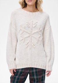 Kobieta ubrana w kremowy sweter z dzianiny z wypukłym wzorem płatka śniegu oraz ciemnozielone spodnie w kratę, stojąca na jednolitym białym tle.