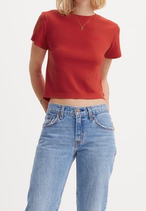 Femme portant un t-shirt rouge Levi's raccourci et un jean en denim bleu clair taille haute, avec un collier en or et des tatouages sur les bras.