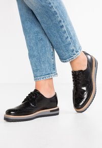 Chaussures à lacets en cuir verni noir avec des détails en brogue et une semelle contrastante beige et grise, portées avec un jean en denim bleu clair retroussé.