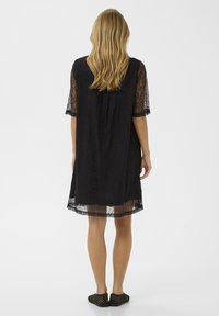 Sieviete ar gariem blondiem matiem, ģērbusies melnā mežģīņu kleitā līdz ceļiem ar caurredzamām piedurknēm un melnām austām čībām, stāv ar muguru vērsta.