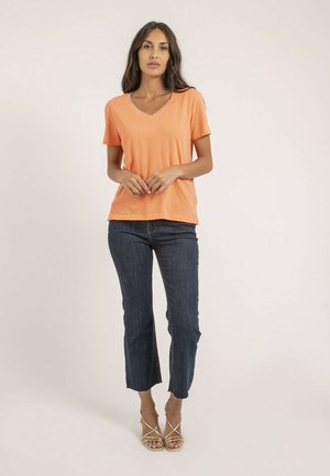 T-shirt orange à col en V à manches courtes, coupe décontractée, associé à un jean cropped bleu marine et des escarpins beige à lanières, se tenant devant un fond neutre.