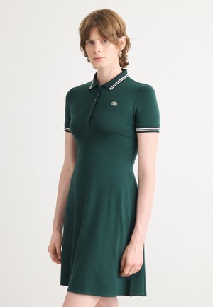 Abito polo verde scuro con maniche corte, colletto e polsini a righe bianche, e un piccolo logo ricamato sul lato sinistro del petto. Tessuto liscio.