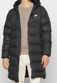 Svart puffjacka med dragkedja framtill, justerbar huva och sidofickor. Har ett quiltat mönster och en liten vit logotyp på bröstet.