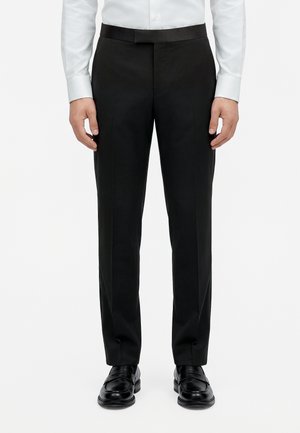 Une personne se tient devant un fond blanc uni, vêtue d’un pantalon noir ajusté, d’une chemise blanche et de mocassins en cuir noirs.