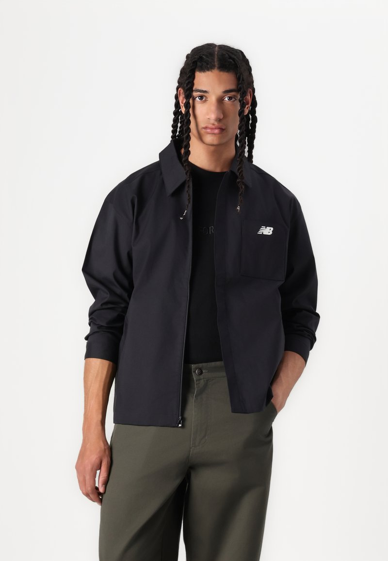 Veste noire légère avec un col, fermeture éclair et un logo brodé sur la poche de poitrine. Portée sur une chemise noire et un pantalon olive.
