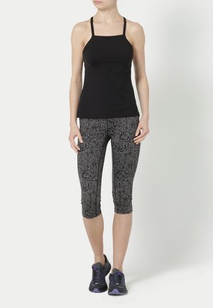 Kvinde iført sort ærmeløs top, mønstrede grå capri leggings og sorte løbesko med lilla detaljer, stående mod en ensfarvet baggrund.