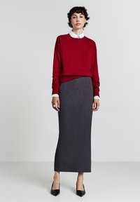 Maglione di cotone rosso sopra una gonna a matita lunga grigia, con una camicia bianca con colletto sotto. Tacchi a punta neri e anelli d'argento sulle mani.