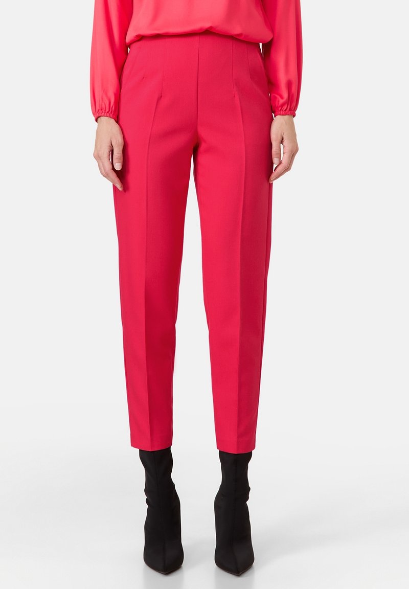 Vrouw in hoogge tailleerde, op maat gemaakte felrode broek met centrale plooien en zwarte puntige enkelboots, staande tegen een effen achtergrond.
