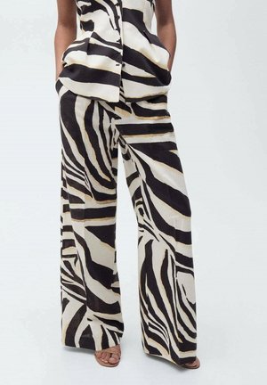 Frau trägt passende schwarz-weiß-beige zebragestreifte weit geschnittene Hose und ärmelloses, geknöpftes Oberteil mit Händen in den Taschen.