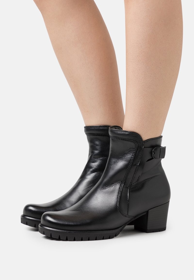 Bottines en cuir noir à talons blocs et boucle latérale portées sur jambes nues sur un fond clair.
