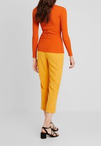 Top de manga larga naranja con canalé combinado con pantalones amarillos cortos y sandalias de tacón negro con tiras, visto desde atrás.