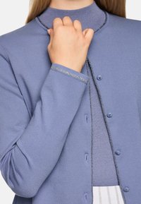 Leichter blauer Strickcardigan mit silberner Bordüre, rundem Halsausschnitt und Knopfleiste vorne. Ärmel mit leichter Textur und figurbetontem Schnitt.