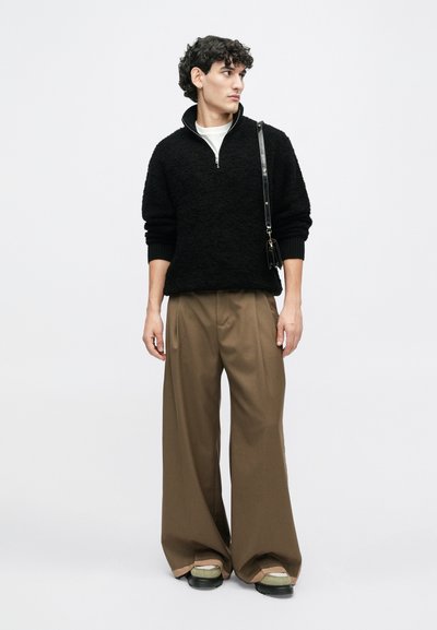 Schwarzer strukturiert Pullover mit hohem Kragen, kombiniert mit weiten braunen Hosen. Das Modell trägt grüne und schwarze Schuhe und hat eine kleine schwarze Tasche dabei.