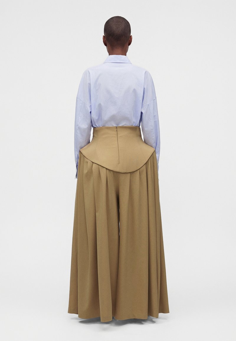 Personne portant une chemise bleu pâle et un pantalon large camel taille haute avec des hanches structurées et évasées, debout de dos sur un fond blanc.