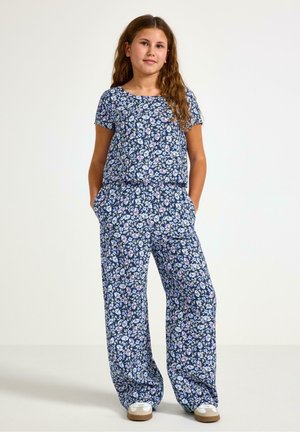 Dívka stojí s rukama v kapsách, má na sobě modrý květovaný krátký rukáv a široké kalhoty, doplněné bílými teniskami.