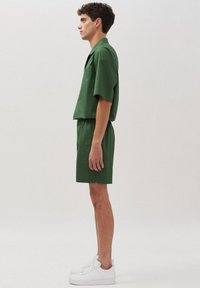 Set di camicia verde a maniche corte e pantaloni abbinati, realizzato in tessuto morbido, con tasca sul petto e fessure laterali. Indossato con scarpe da ginnastica bianche.