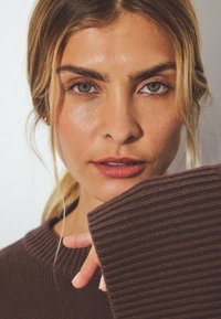 Brun ribbad tröja med en vid halsringning, närbild av ansiktet som visar gröna ögon och mjuka rosa läppar, hår stylat i lösa vågor.