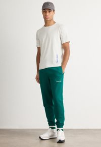 Svetlo šedé tričko s modrým logom Calvin Klein, spárované so zelenými teplákmi s logom Calvin Klein, biele tenisky s čiernymi podrážkami.