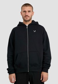 Felpa nera con zip in cotone, dotata di tasca a marsupio, polsini a coste e una piccola grafica bianca sul lato superiore sinistro del petto.