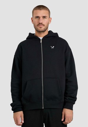 Musta vetoketjullinen huppari, joka on valmistettu puuvillasta. Siinä on kengurupocket, ribbaus hihansuissa ja pieni valkoinen graafinen kuvio vasemmassa rintakehässä.