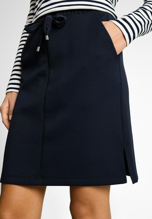 Frau trägt einen marineblauen Rock mit seitlichem Schlitz und Kordelzugbund, Hand in der Tasche, kombiniert mit einem schwarz-weiß gestreiften Langarmshirt.