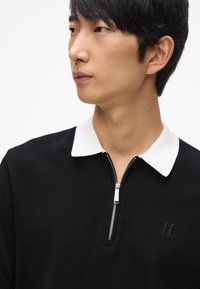 Crni pulover s bijelim patentnim ovratnikom, s prednjim patentnim zatvaračem i izvezenim logotipom, od teksturirane pletene tkanine.