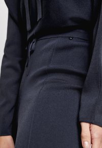 Svart, stickad tyg med en slät yta. Har ett dragsko i midjan och ribbade detaljer längs toppen och sidan. Närbild.