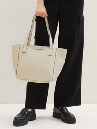 Beige Tragetasche mit strukturiertem geometrischen Muster, schwarzem Rand und doppelten Griffen. Von einer Person gehalten, die schwarze Hosen und Schuhe trägt.