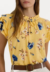 Gul blommig blus med volangärmar, knytning i nyckelhålet och mönster av blå och rosa blommor, parat med ett brunt bälte.